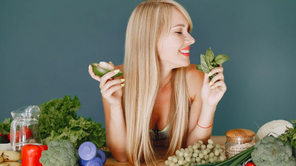 A smiling woman holding avocado and basil, surrounded by fresh vegetables and fruits for healthy eating.